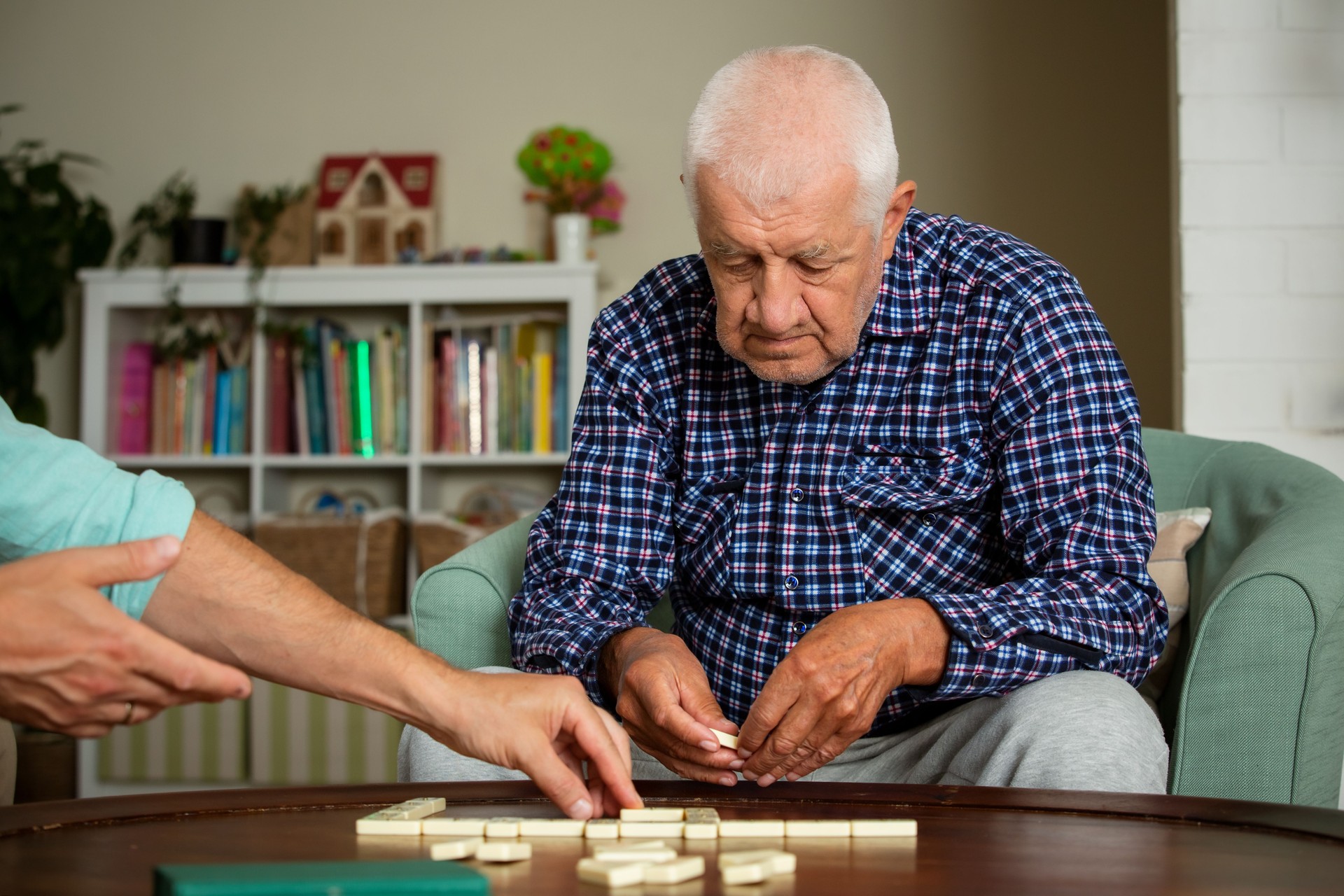 A grown son and his elderly father enjoy a quiet game of dominoes in the living room, sharing connection, laughter, and meaningful time together.